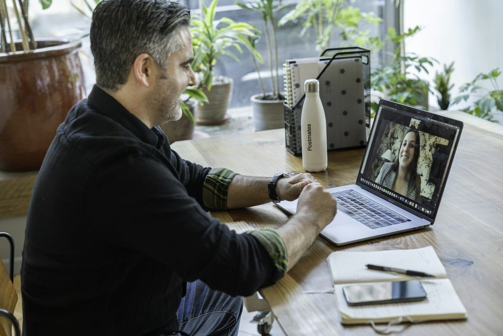 A man on a video call at his desk, a notebook beside the laptop